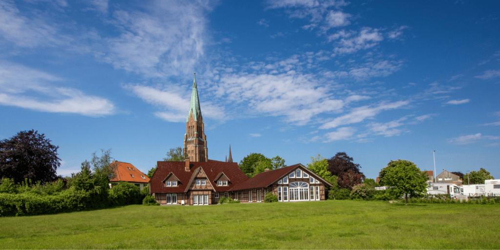 Dağınık bulutların olduğu mavi gökyüzünün altında küçük bir evin arkasında, Schleswig-Holstein'da yüksek bir kulesi olan geleneksel bir kilise.