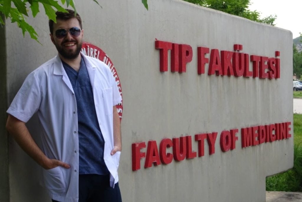 Üzerinde "Benim Maceram Tıp Fakültesi" yazan bir duvarın önünde beyaz önlüklü bir adam duruyor.