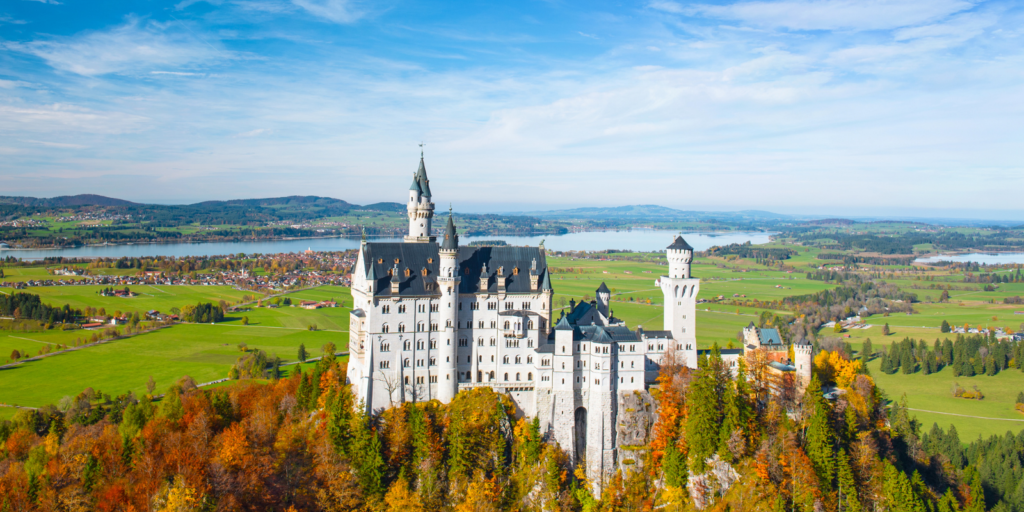 Bayern'de ormanlık bir tepenin üzerinde yer alan, uzakta bir göl bulunan doğal bir manzaraya bakan masalsı bir kale.