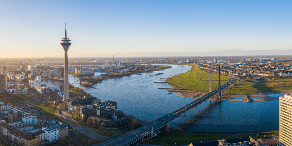 Nordrhein-Westfalen'de bir nehrin yanında belirgin bir kule ve gün batımında köprüler bulunan bir şehir silüetinin havadan görünümü.